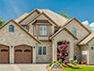 Elegant two-story house with beige stone exterior, brown roof shingles, and two wooden garage doors, surrounded by a well-manicured lawn and colorful flowerbeds under a blue sky with fluffy clouds